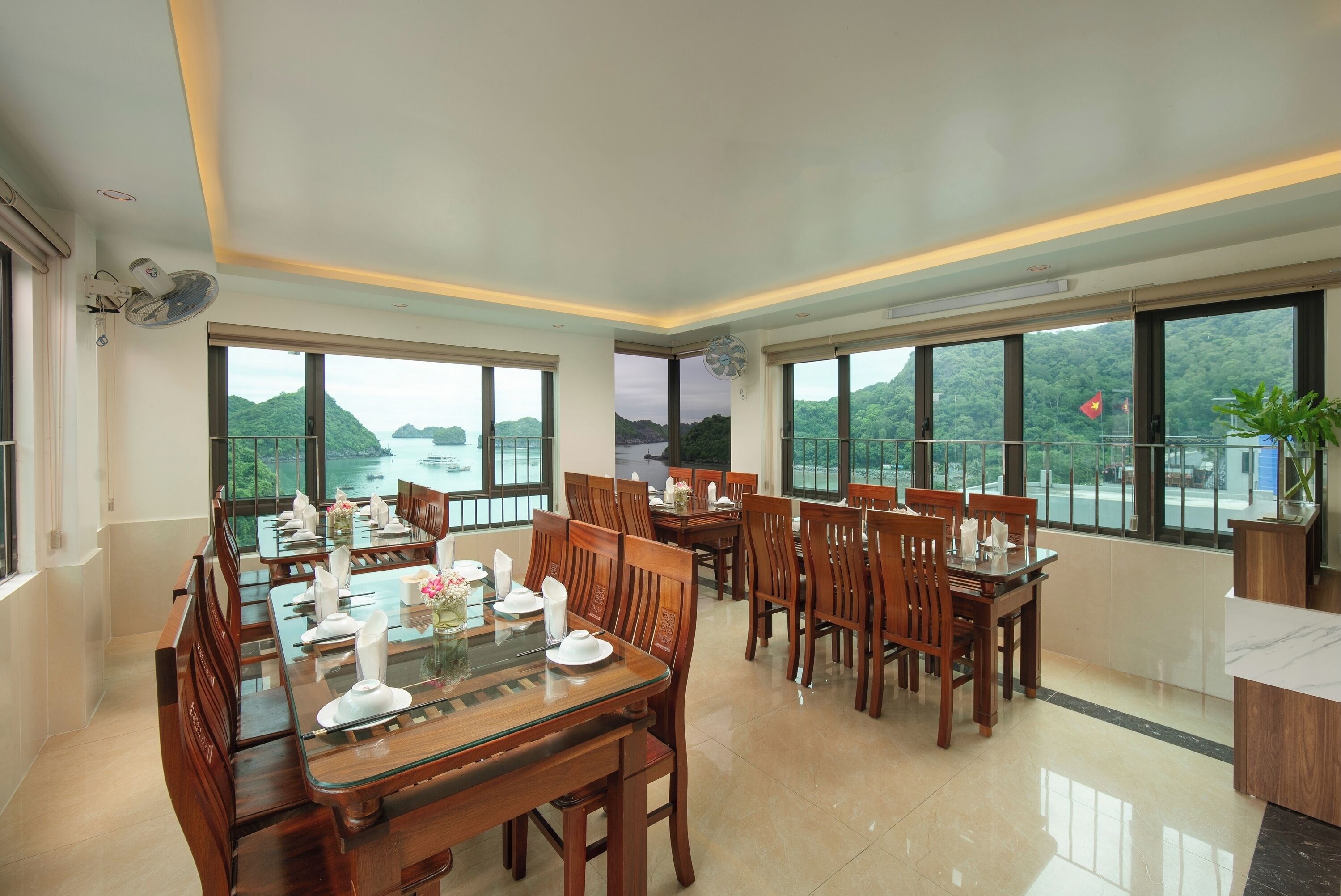 A dining area with multiple tables and chairs, large windows with a view of the sea and hills, and ceiling fans.