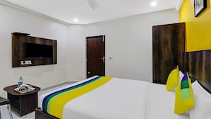 Quarto de hotel com uma cama, uma televisão fixada em um painel de madeira, uma porta e uma estante na parede.