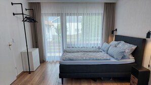 Ett modernt sovrum med en stor säng, ett sängbord, en lampa och ett fönster med tunna, genomskinliga gardiner.