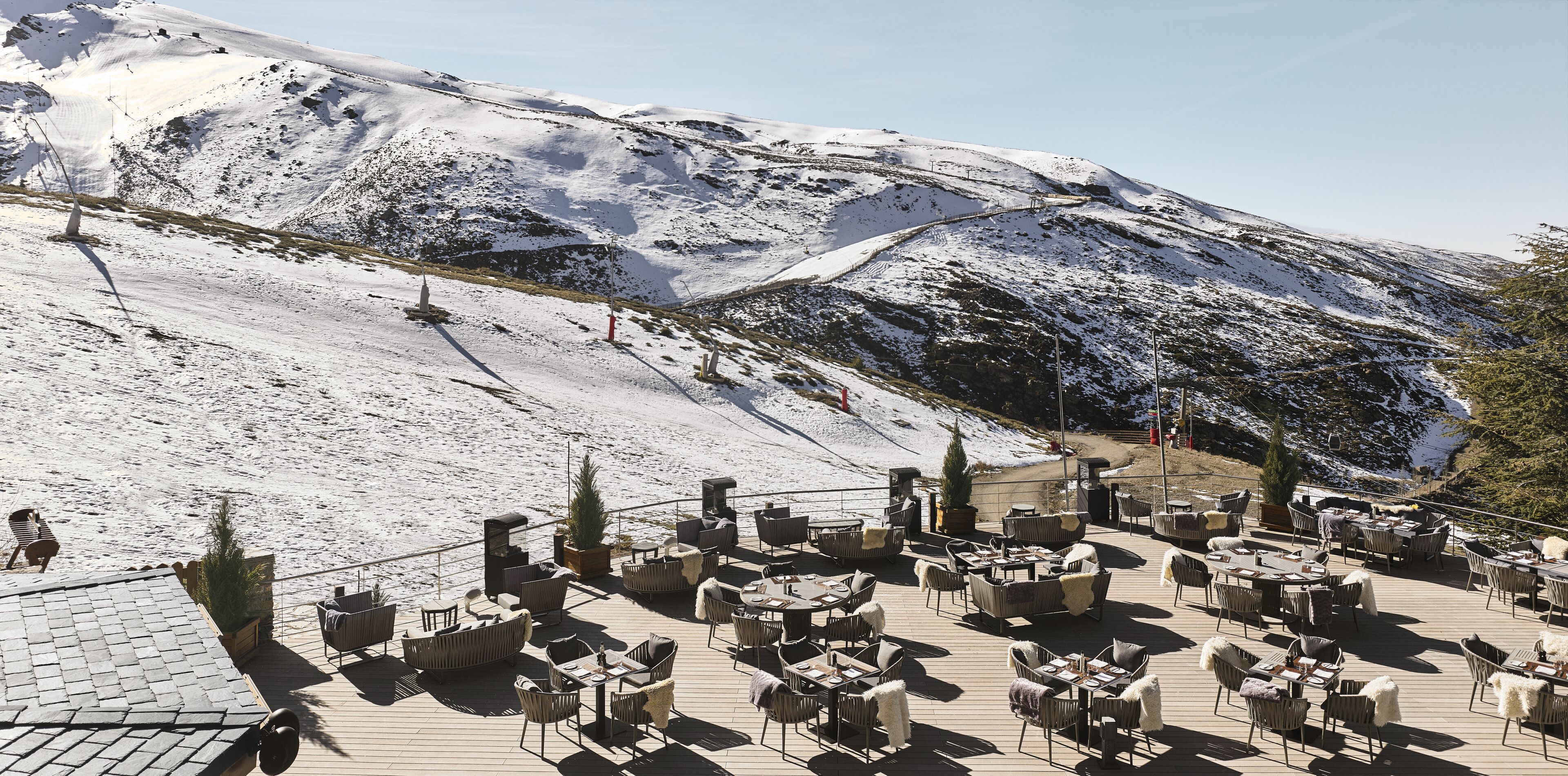 A snow-covered mountain with a ski lift in the background. In the foreground, there is a wooden deck with outdoor seating and tables.