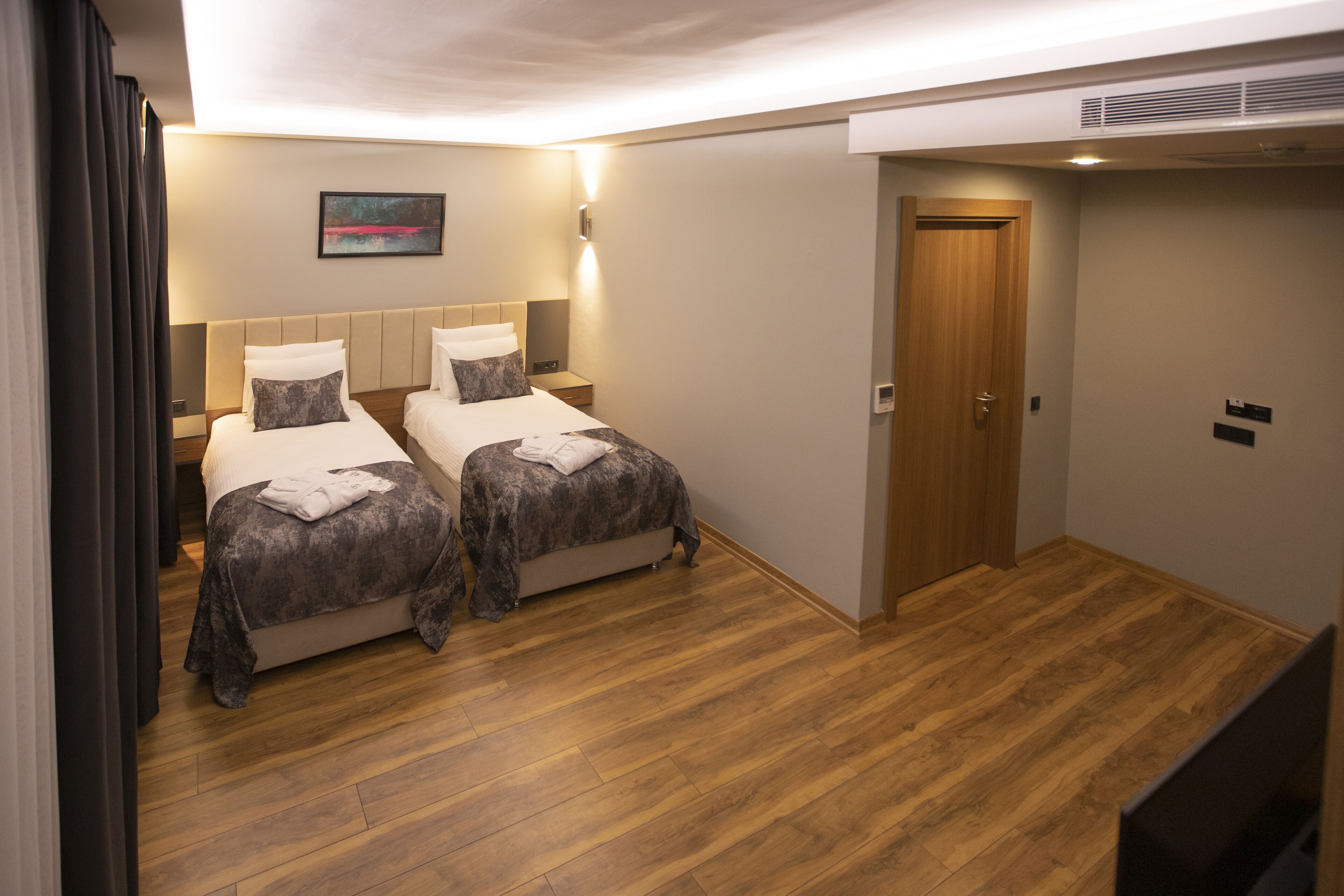 A hotel room with two beds, wooden flooring, a door, and a wall-mounted light fixture.