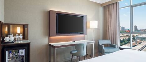 Quarto de hotel com cama, escrivaninha com televisão, cadeira, minibar e uma grande janela com vista para a cidade.
