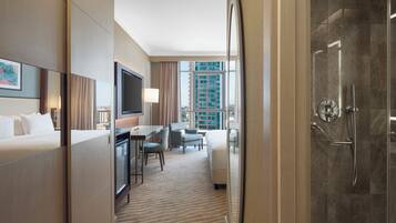 A hotel room with a bed, desk, chairs, a TV, and a shower area.