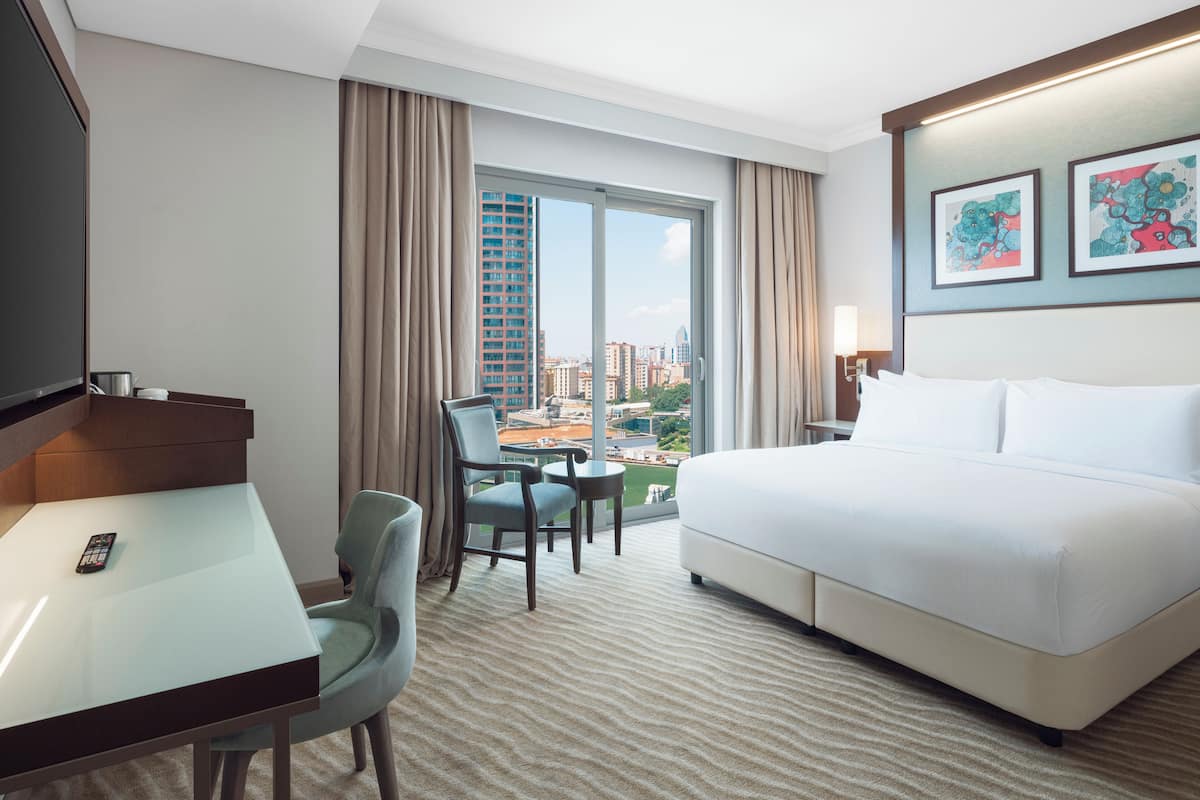 A hotel room with a bed, a desk, a chair, a TV, and a window with a city view.