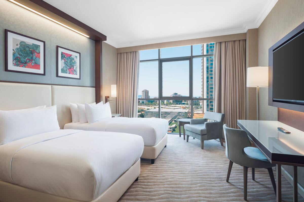 A hotel room with two beds, a desk, a chair, a TV, and a large window with a city view.