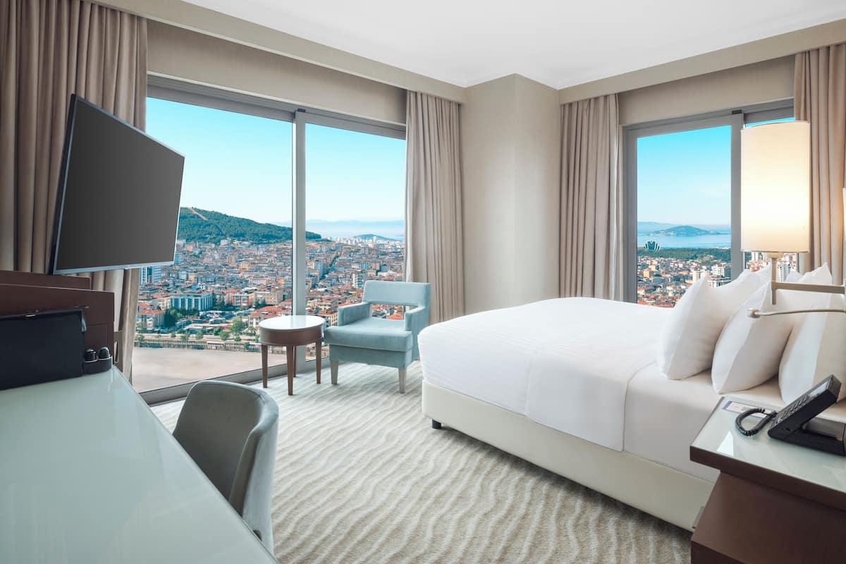 A hotel room with a large bed, a desk with a telephone, a chair, and a television. The room has large windows offering a city view.