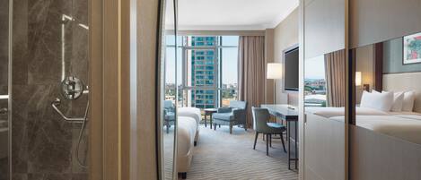 A hotel room with a shower, a bed, a desk, and a view of the city.