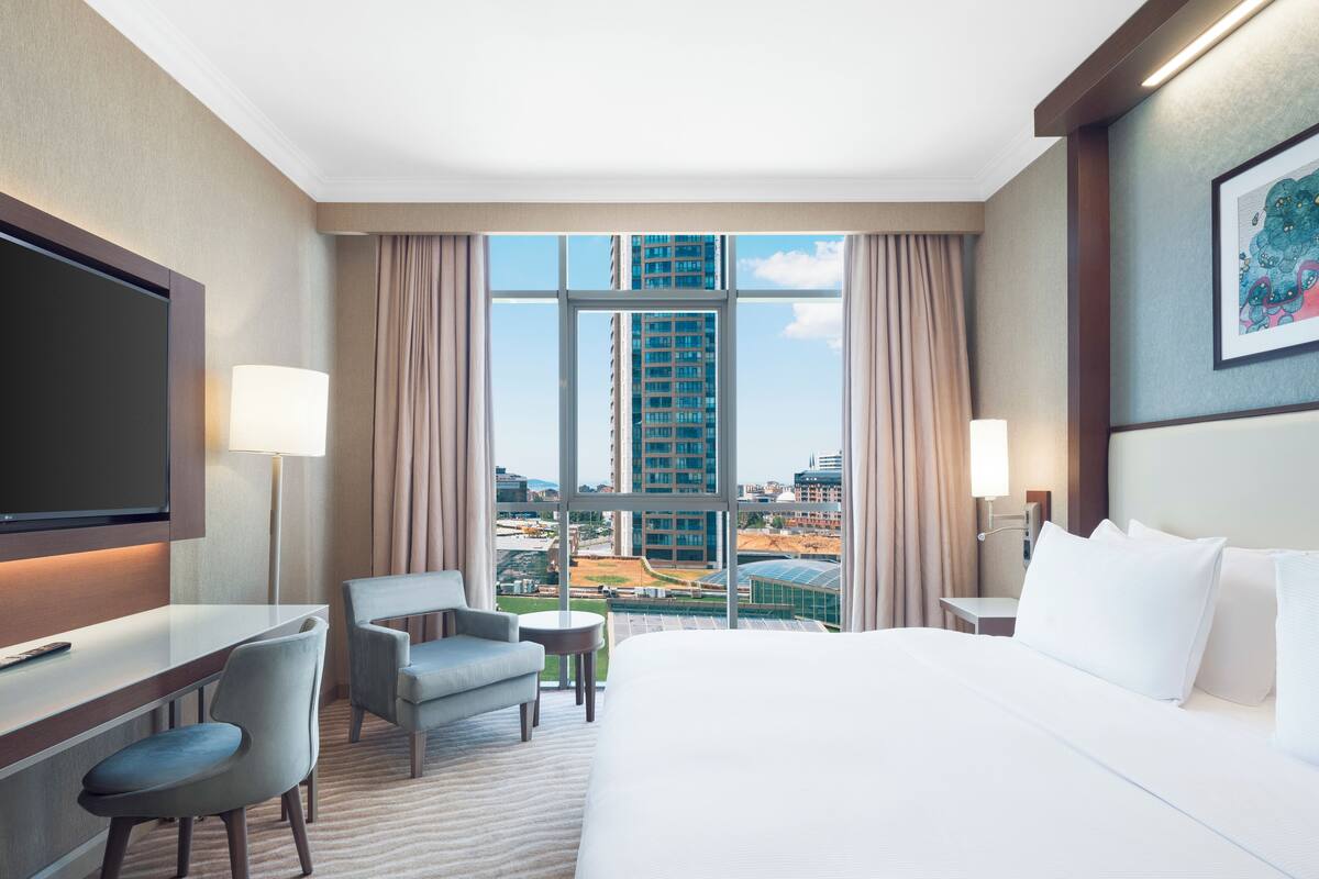 A hotel room with a large bed, a desk, a chair, a TV, and a view of the cityscape through large windows.
