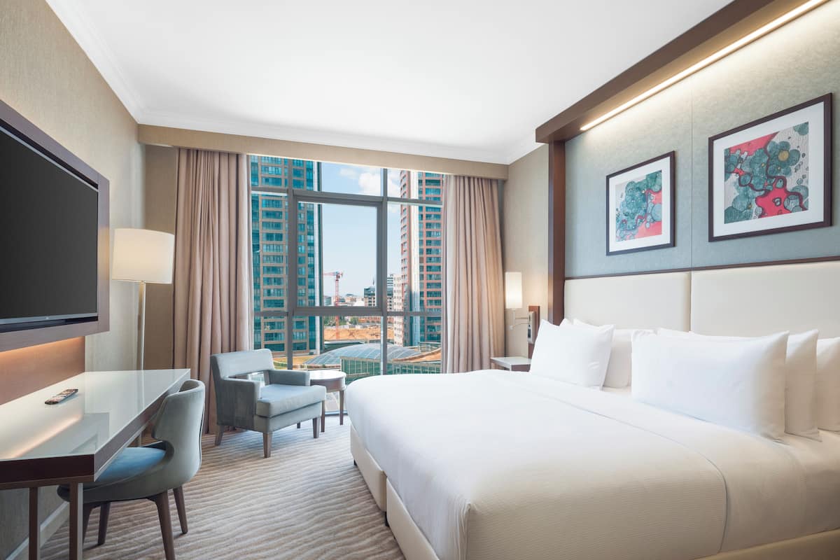 A hotel room with a large bed, a desk with a chair, a TV, and a view of tall buildings.