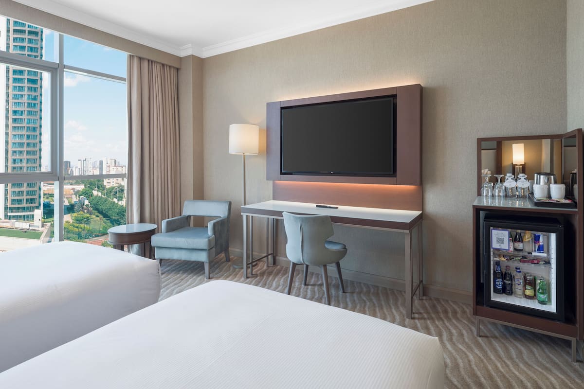 A hotel room with two beds, a desk with a flat-screen TV, a minibar, and a large window with a city view.