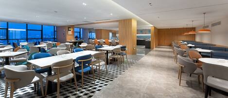 A modern dining area with multiple tables and chairs, a check-in counter, and large windows offering city views.