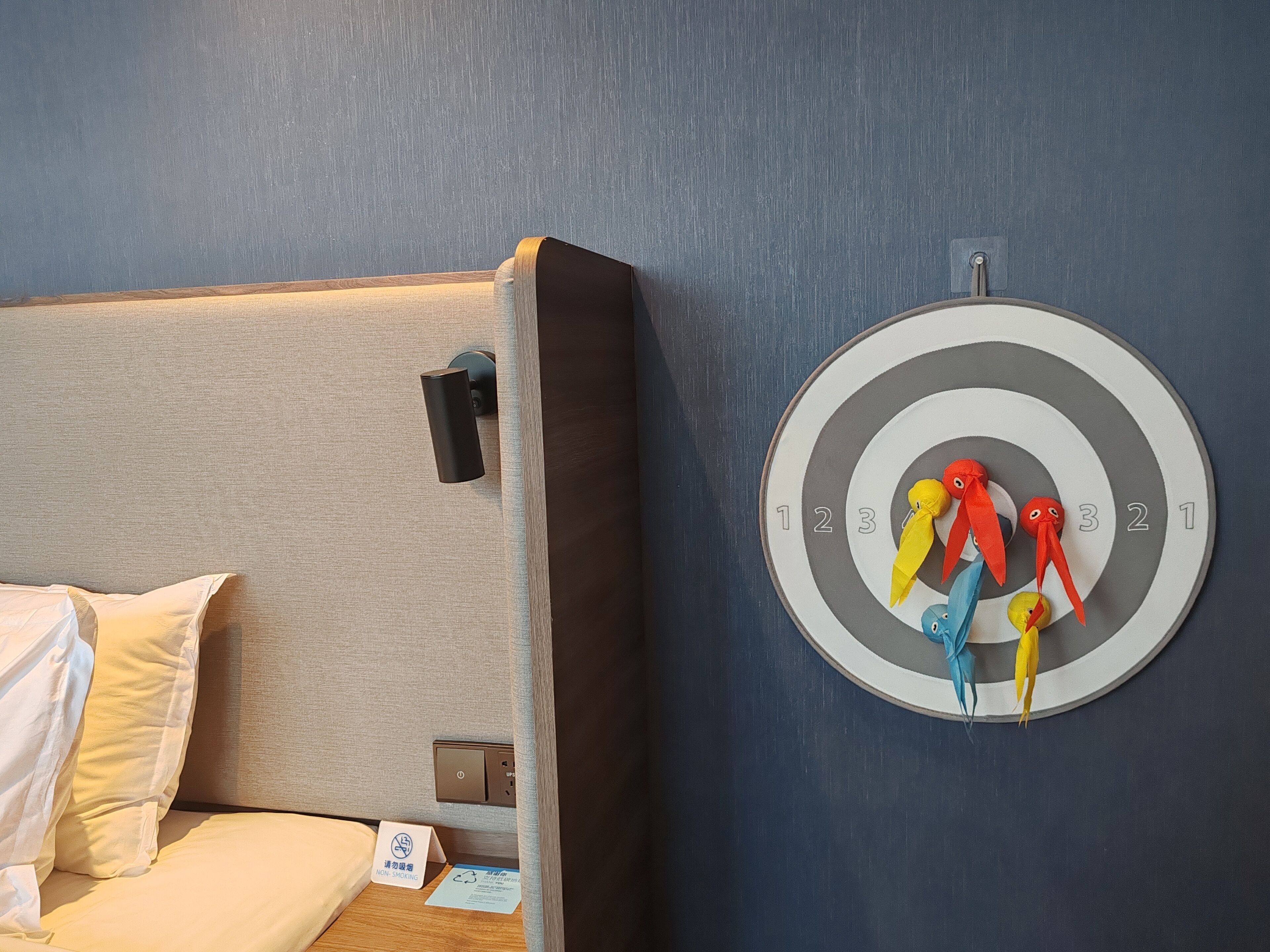 A hotel room with a bed, a wall-mounted dartboard, and a non-smoking sign.