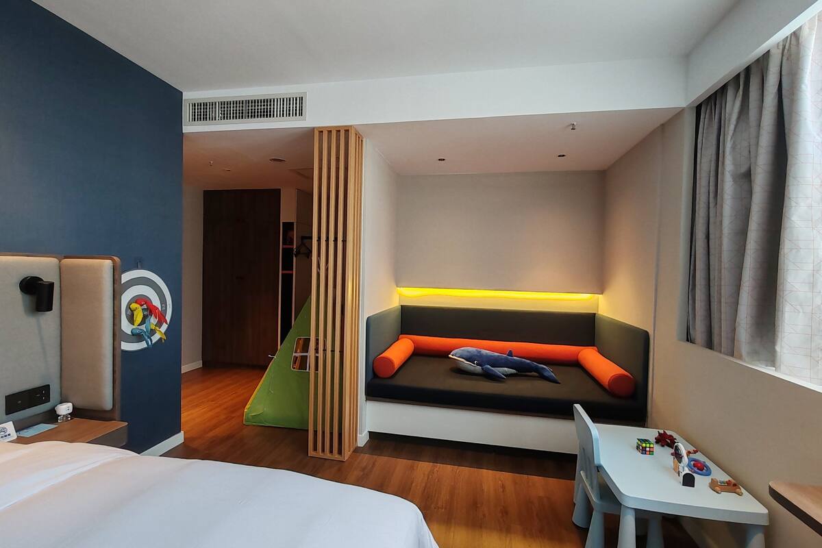 A hotel room with a bed, a desk with toys, a colorful wall mural, and a seating area with cushions.