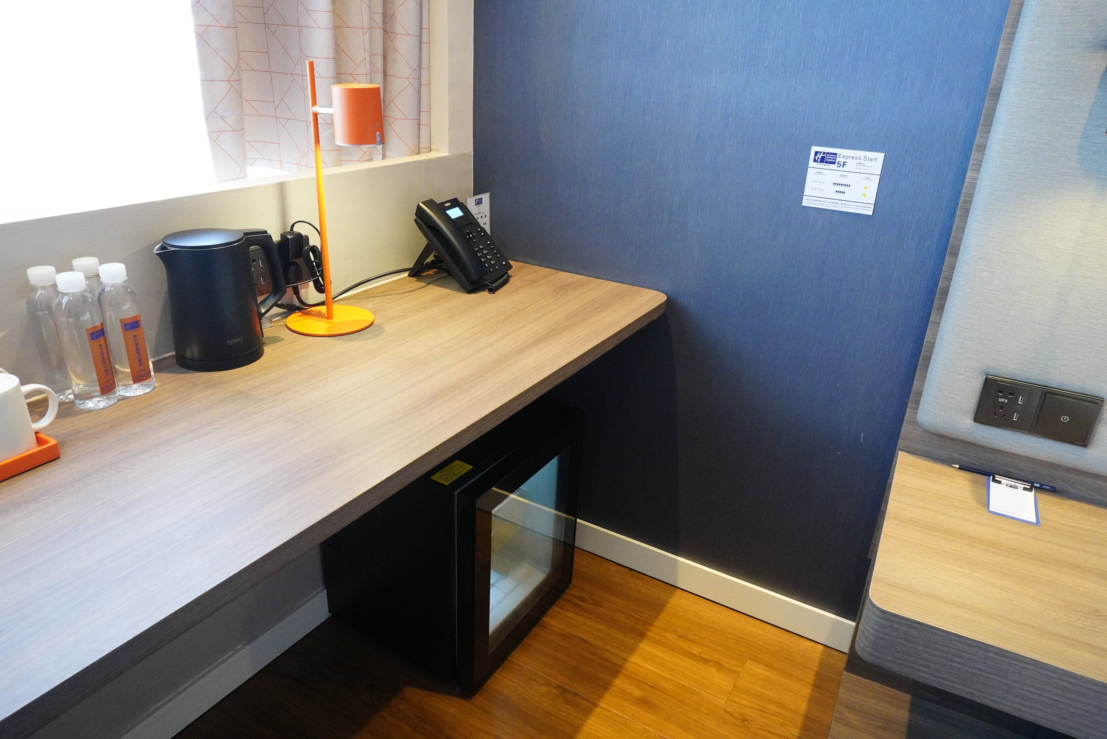 A hotel room with a wooden desk, a coffee maker, a phone, and a small refrigerator.