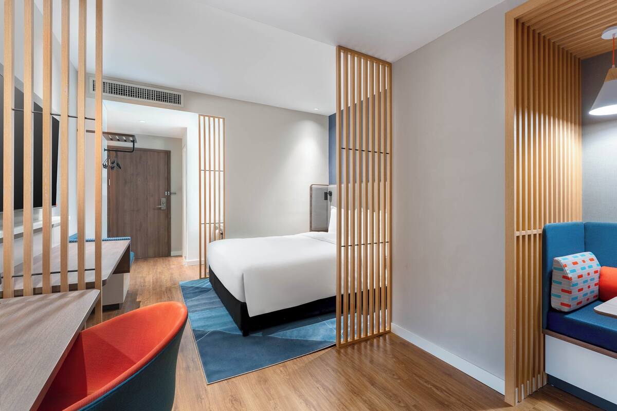 A modern hotel room with a wooden partition, a bed, a blue sofa, a small table with a vase, and a desk with a chair.
