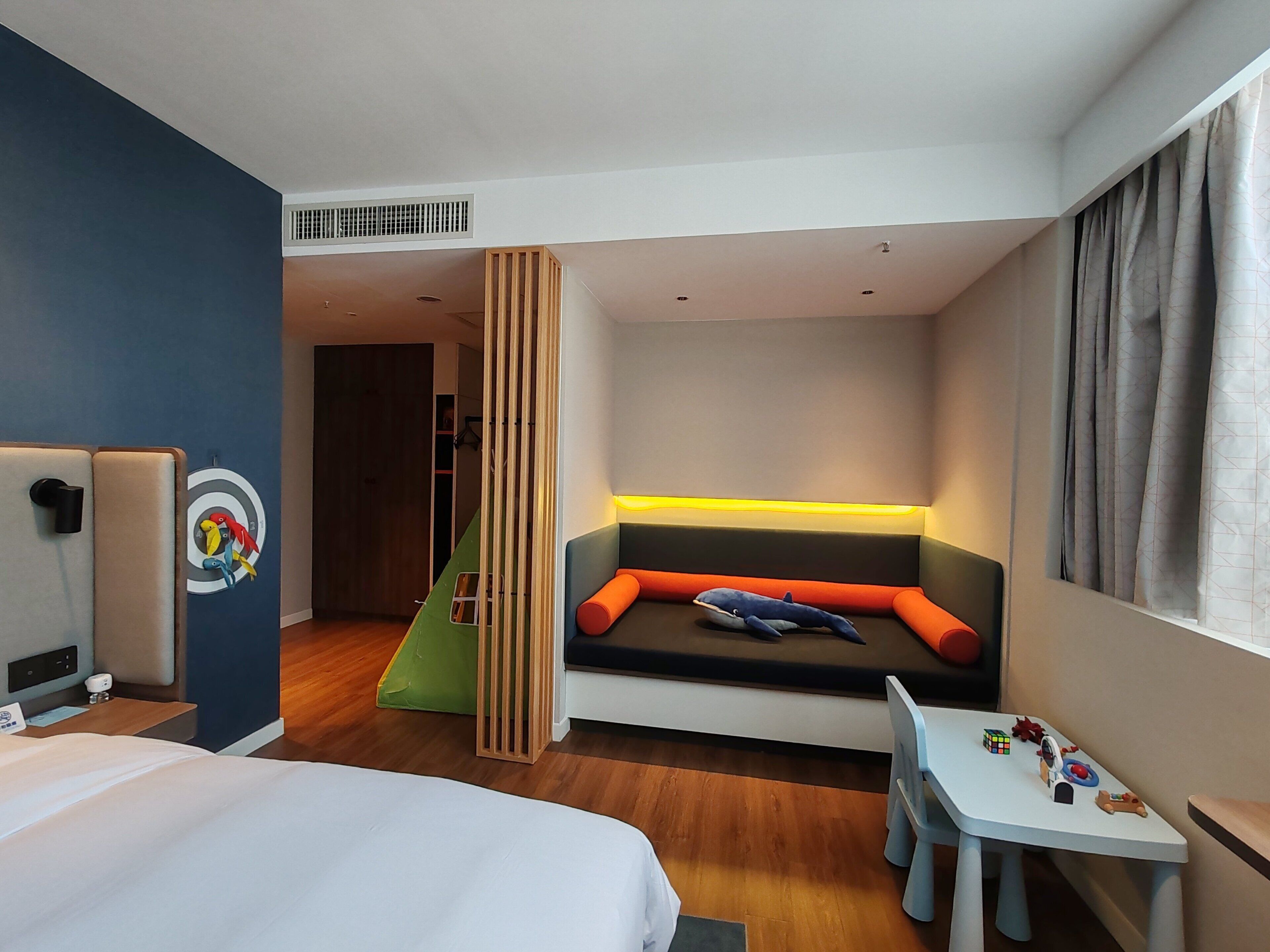 A hotel room with a bed, a desk with toys, a colorful wall mural, and a seating area with cushions.