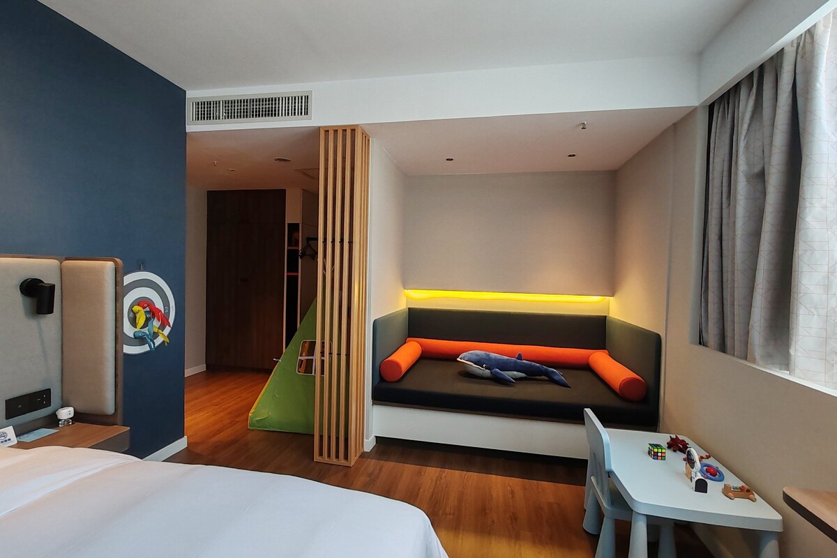 A hotel room with a bed, a desk with toys, a colorful wall mural, and a seating area with cushions.