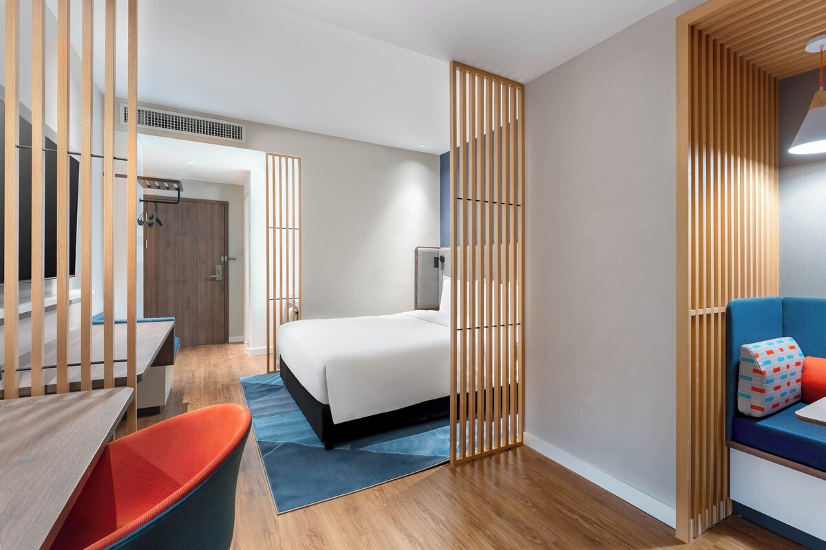 A modern hotel room with a wooden partition, a bed, a blue sofa, a small table with a vase, and a desk with a chair.