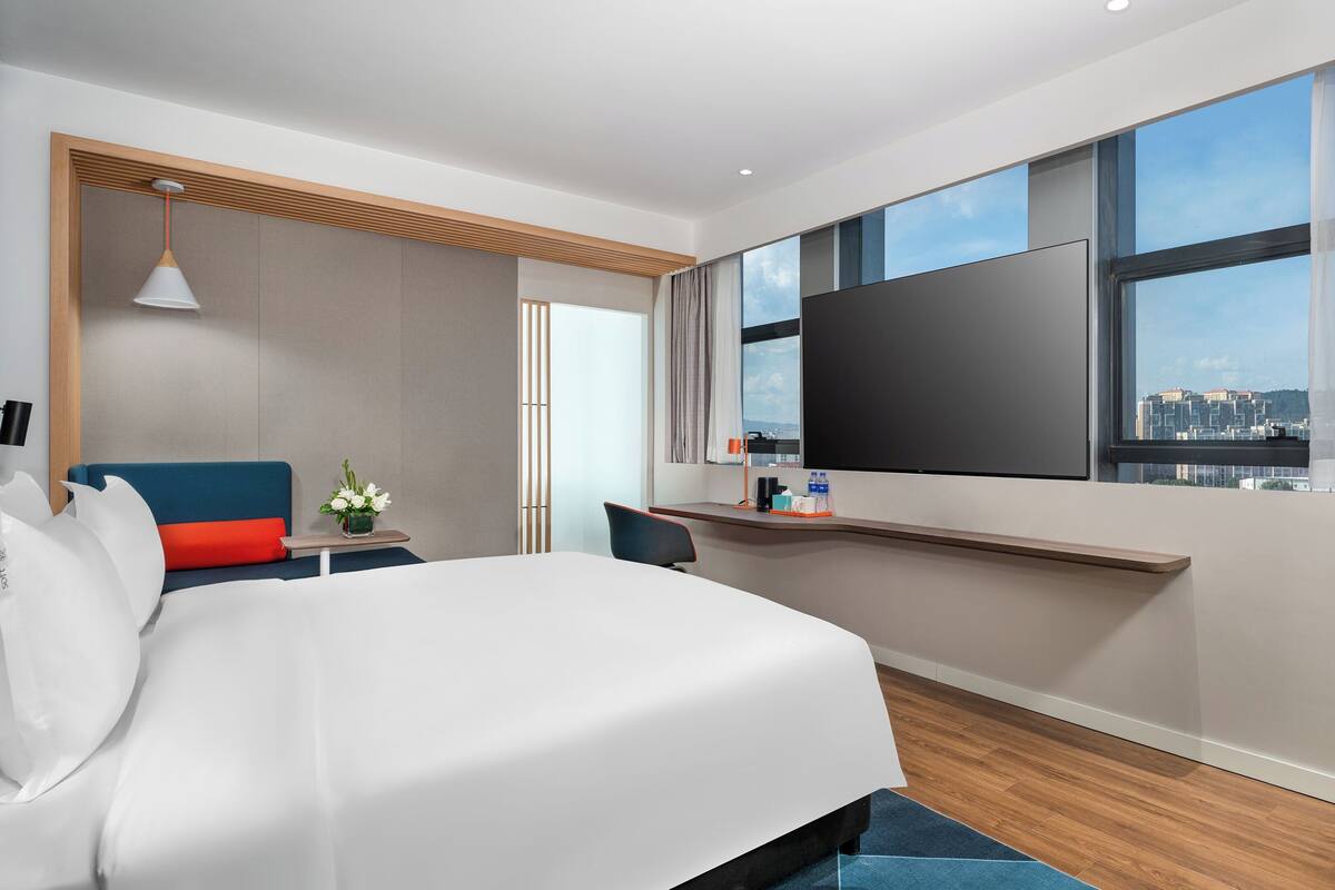 A modern hotel room with a large bed, a desk, a flat-screen TV, and a city view.