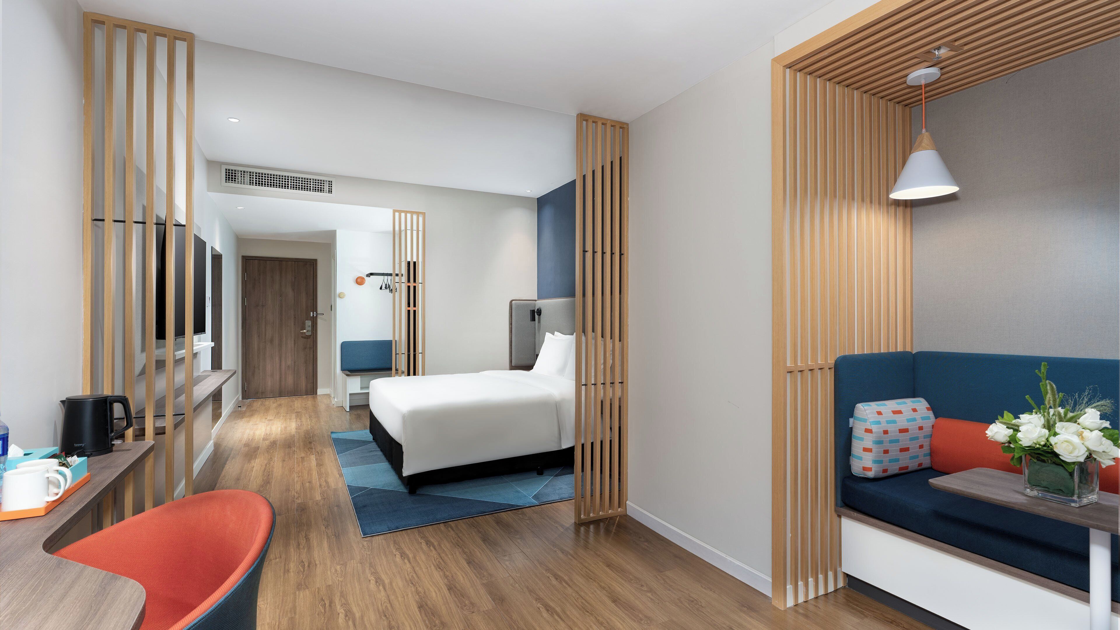 A modern hotel room with a wooden floor, a bed, a desk with a chair, a sofa, and a small table with a vase of flowers.
