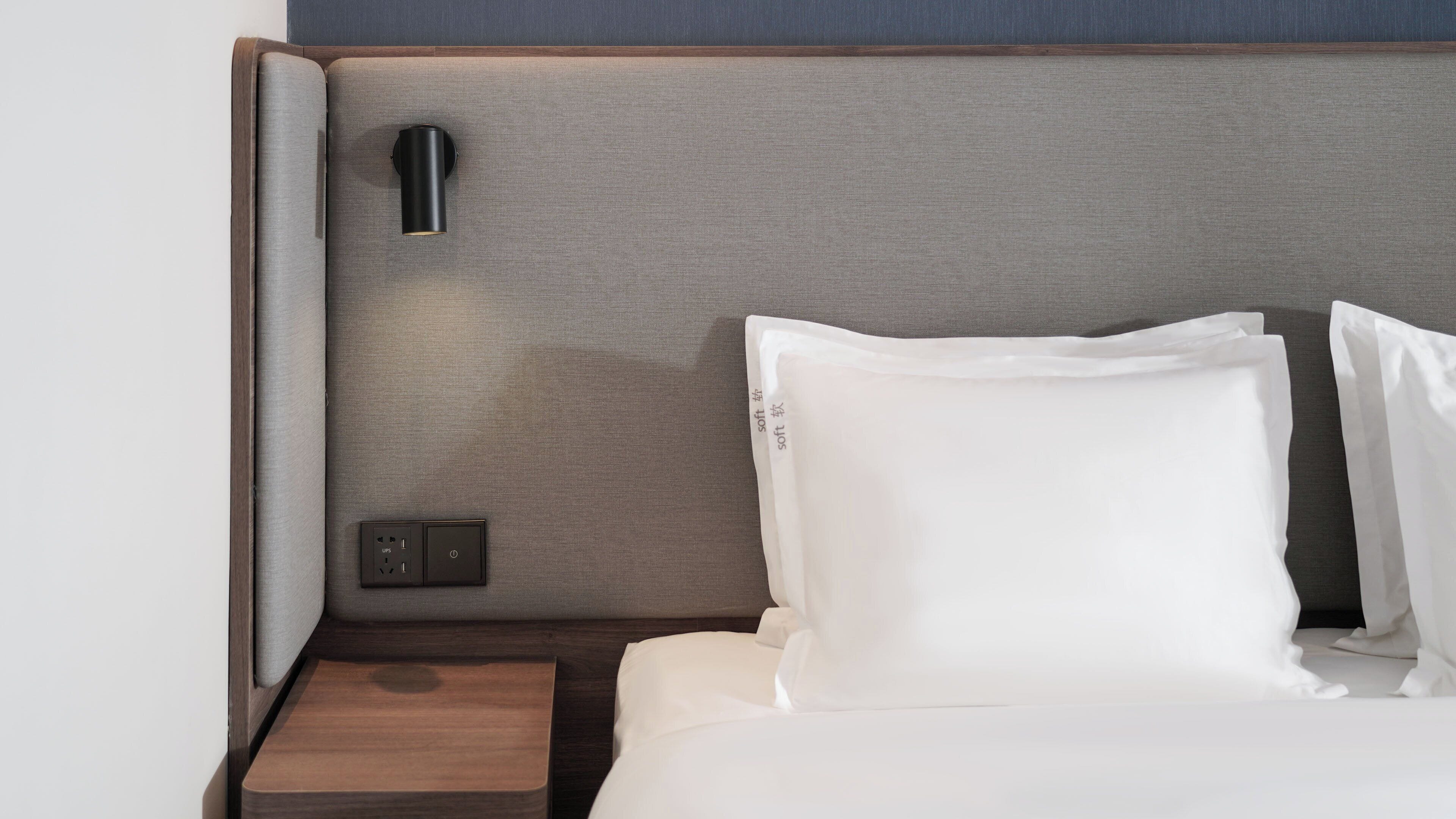 A neatly made bed with white linens, a wooden nightstand with a power outlet, and a wall-mounted light fixture.