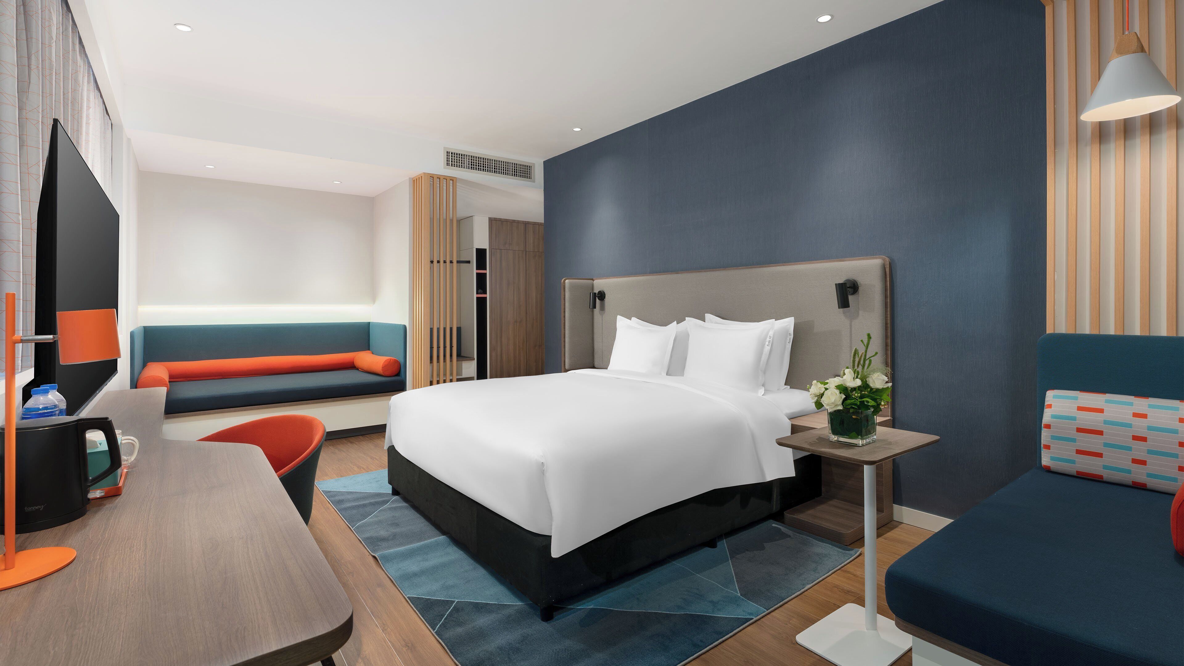 Una habitación de hotel moderna con una cama grande, una zona de estar con sofá y sillón, un escritorio con computadora y una mesita de noche con un florero.