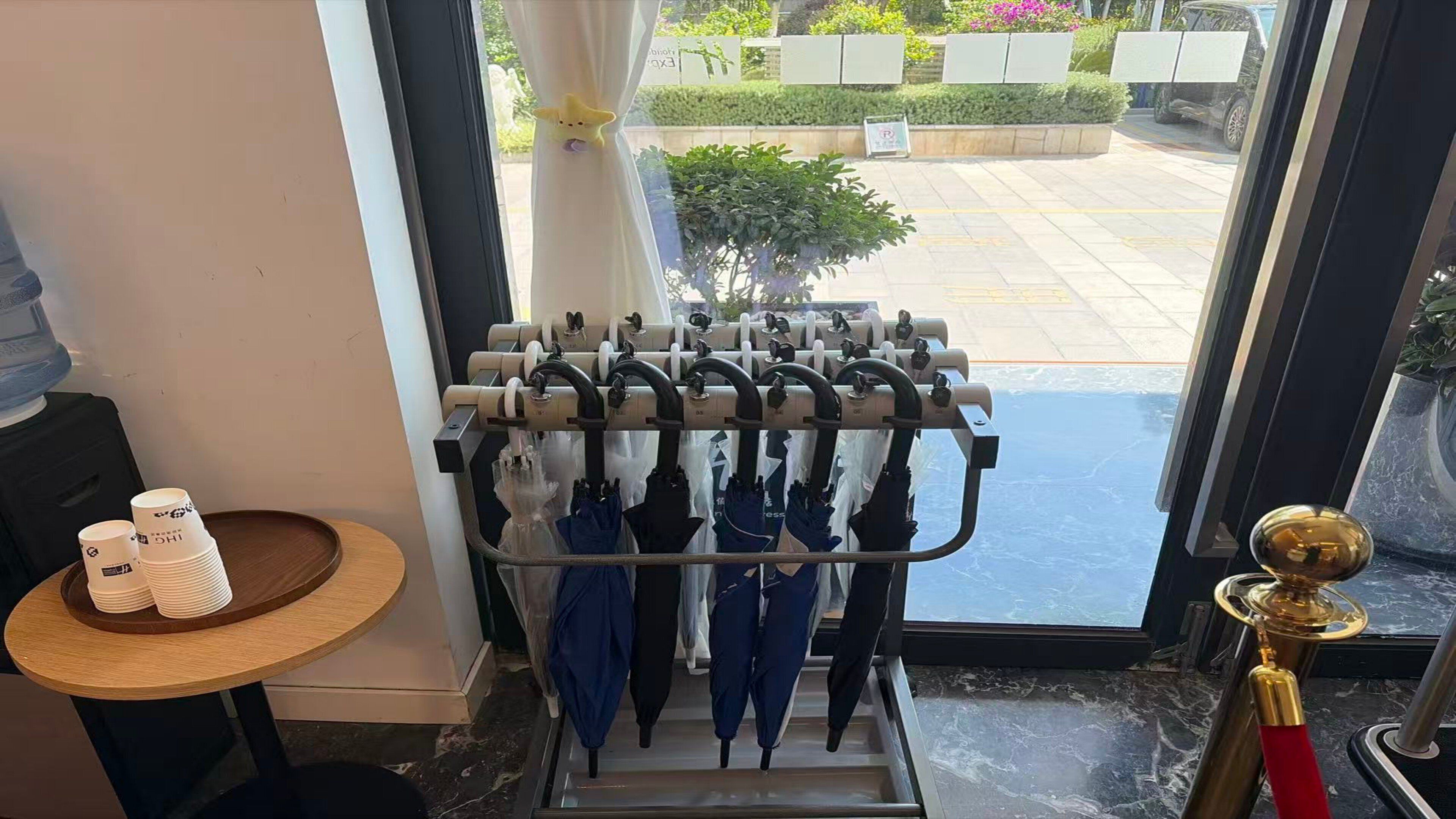 L’entrée d’un hôtel, avec une porte en verre, un porte-parapluies, une petite table sur laquelle sont empilées des assiettes et un distributeur d’eau.