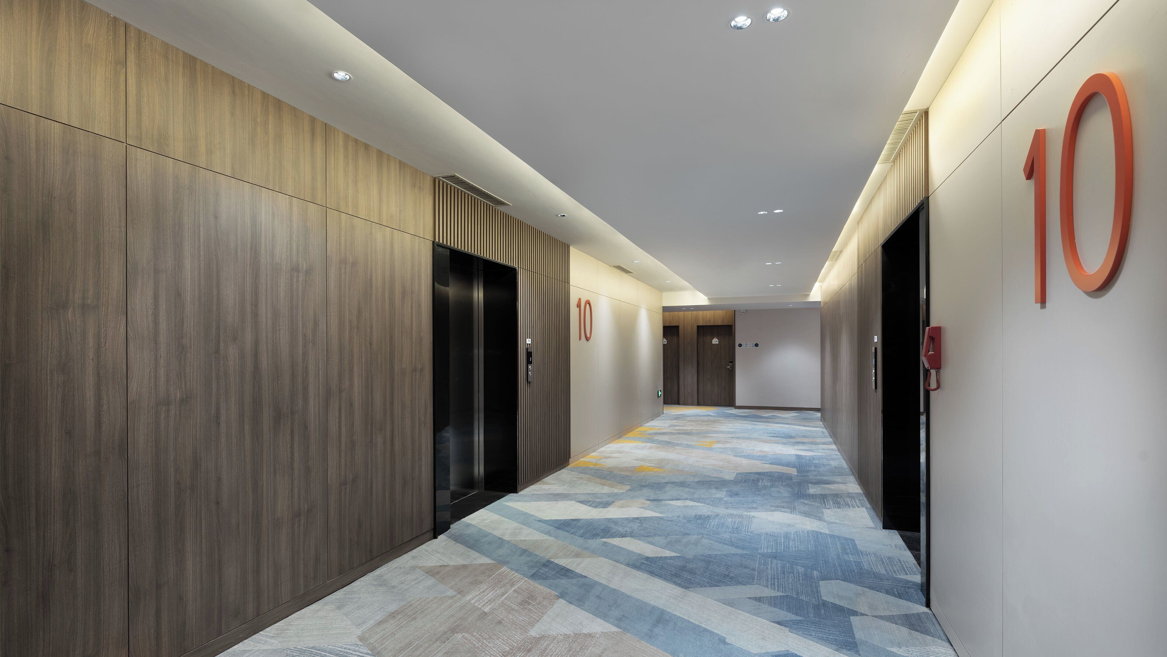 A modern hotel corridor with wooden panel walls, a patterned carpet, and a number '10' sign on the wall.