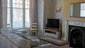 Uma sala de estar com sofá, televisão em um móvel, lareira e janela com cortinas.