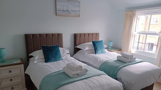Quarto de hotel com duas camas, cada uma com cabeceira, roupa de cama branca, almofadas de lancho verde-água e um cobertor verde-água. Há uma pequena mesa de cabeceira com abajur, uma janela com cortinas e um quadro na parede.