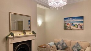 Un salon avec une cheminée, un canapé beige agrémenté de coussins à motifs, un tableau représentant des cabanes de plage et un lustre.