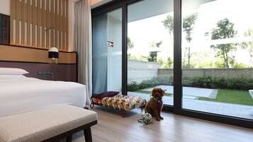 A hotel room with a large bed, a small sofa, a dog, and a view of a garden through a large window.