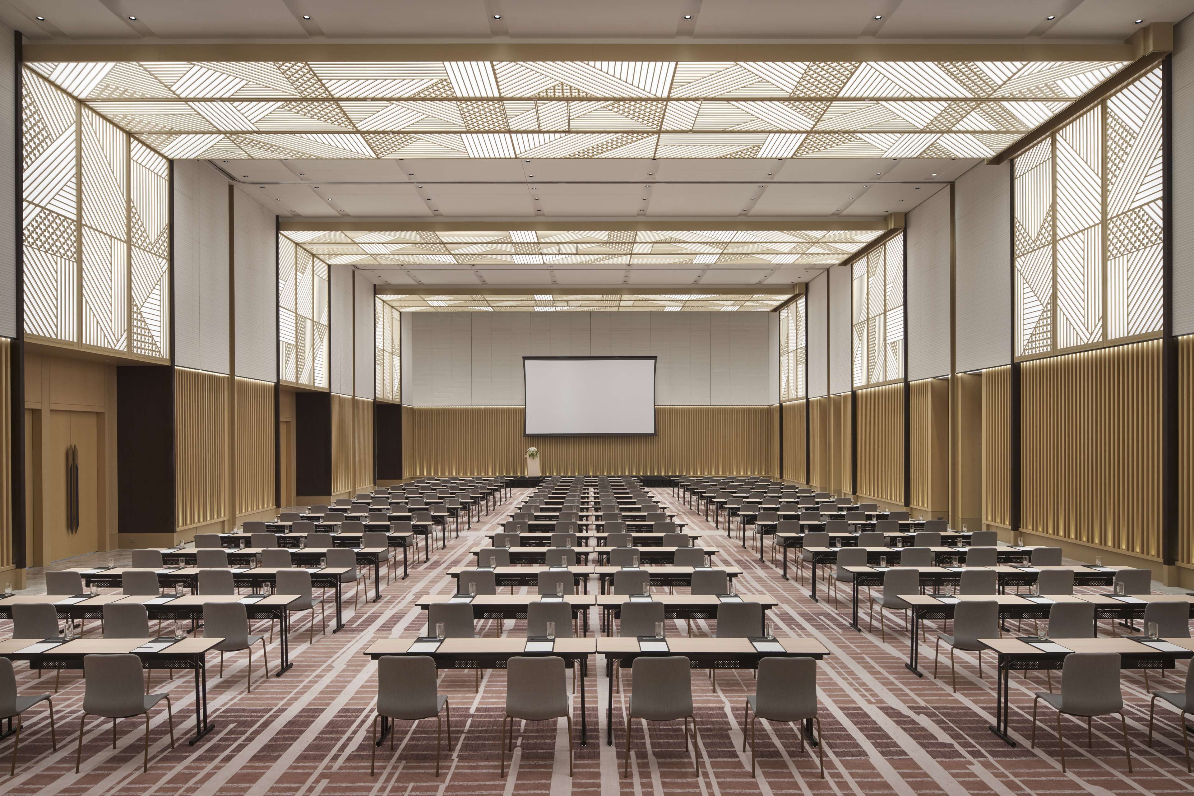 Un salón de conferencias con filas de mesas y sillas, una pantalla grande al frente y paneles de techo estampados.