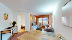 A modern living room with a wooden dining table, a TV, and a sofa. There is a balcony with a view of snow-covered mountains. - The newly renovated holiday apartment "Abitaziun Caspar Badrutt 306" in St. (St. Moritz)