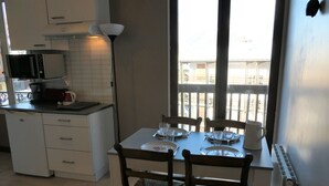 Una cocina compacta con armarios blancos, microondas y cafetera. Hay una mesa de comedor lista con platos, vasos y cubiertos. La habitación tiene un ventanal con vista a un balcón.