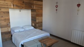 Un dormitorio con cabecera de madera, cama con ropa blanca, un banco gris y un taburete pequeño de madera.