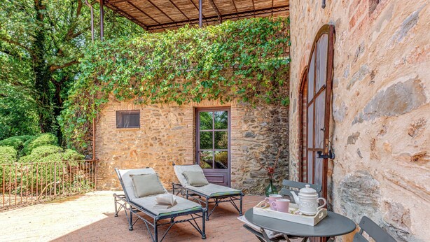 Ein Steinhaus mit einer überdachten Terrasse, zwei Liegestühlen, einem Tisch mit Teekanne und Tassen sowie einem Fenster.