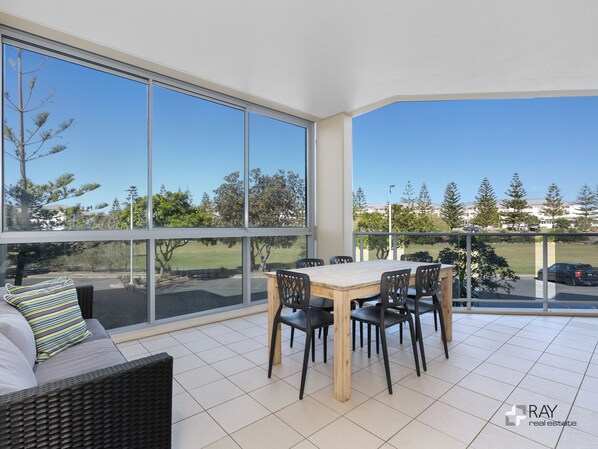A modern living room with a large window, a dining table with chairs, and a sofa with a striped pillow. - Parkview Paradise – 2BR Bali Resort in Kingscliff (Tweed)