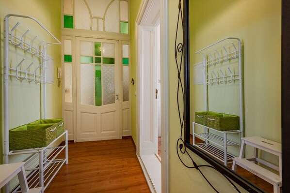 Un pasillo con piso de madera, paredes blancas y una puerta con detalles verdes. Se observan estantes metálicos blancos con cestas en las paredes.