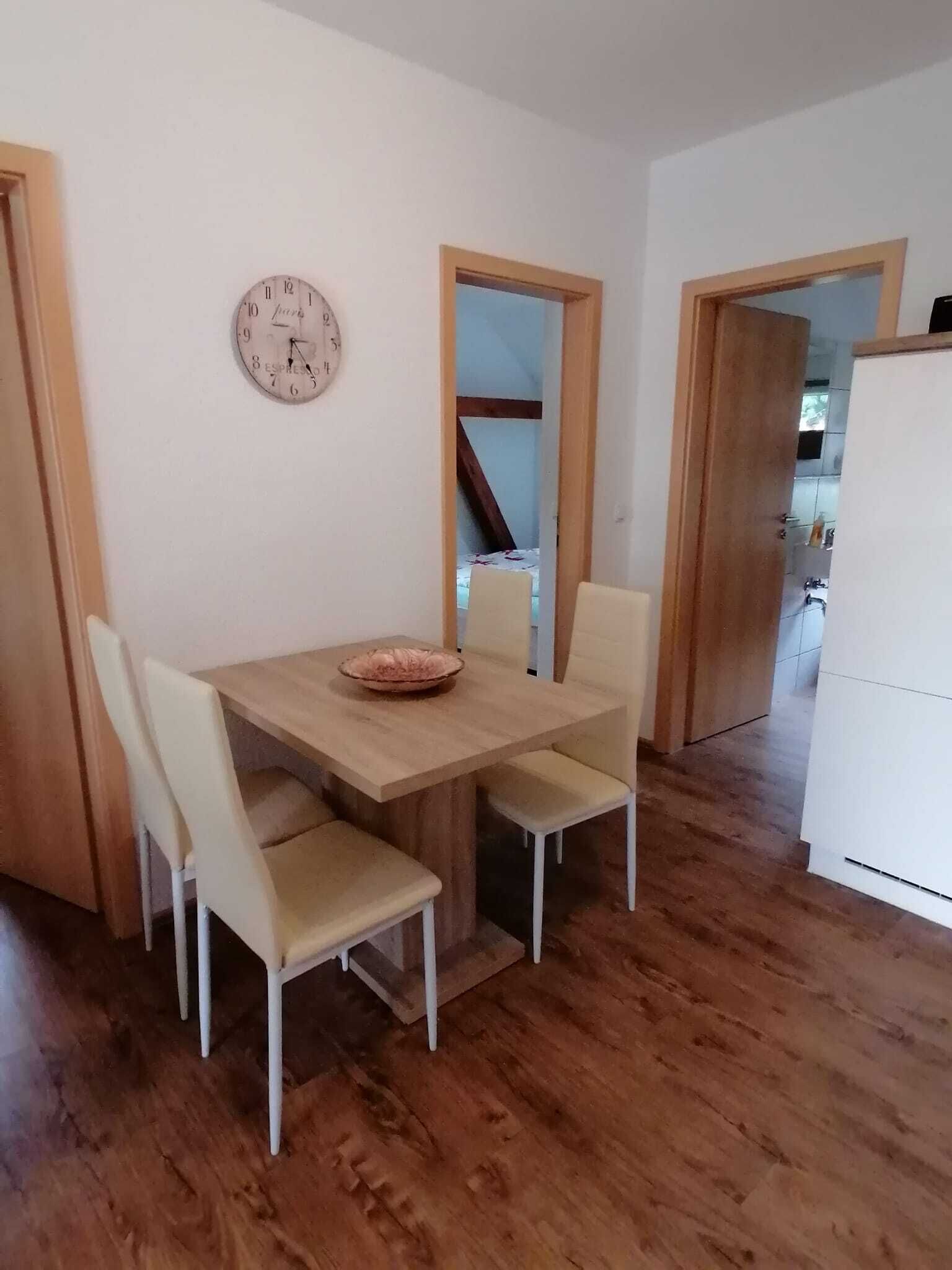 Ein Essbereich mit Holztisch und vier Stühlen, einer Uhr an der Wand und einer durchgehenden Tür erkennbaren Küche.