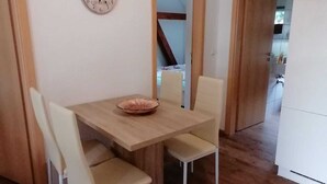 Een eethoek met een houten tafel en vier stoelen, een klok aan de muur, en een keuken die zichtbaar is door een open deur.