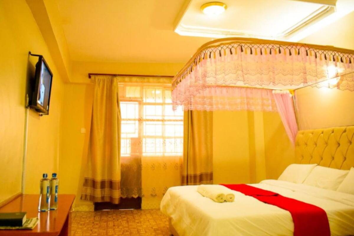 A hotel room with a bed, a television on the wall, a desk with a book and bottles, and a window with curtains.