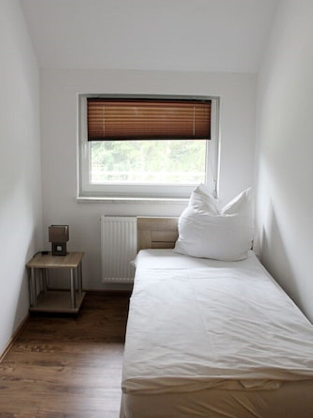 A small, clean room with a single bed, a nightstand, a window with blinds, and a radiator.
