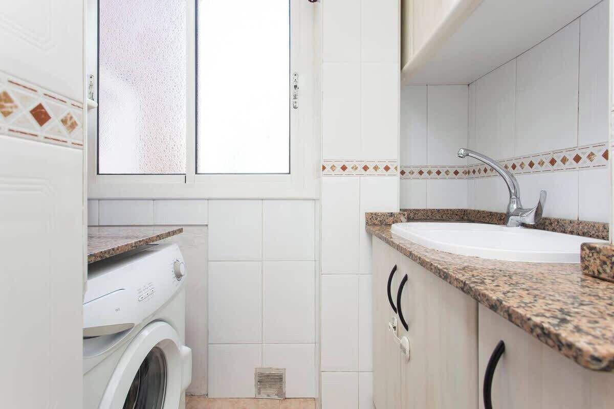 A compact kitchen with a built-in washing machine, a sink with a granite countertop, and white tiled walls.