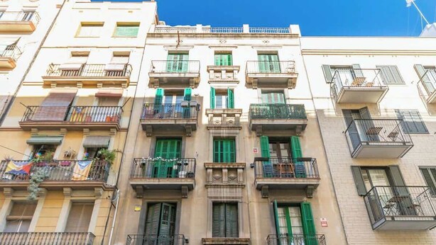 Un immeuble à plusieurs étages, doté de balcons, de volets verts et surplombant un ciel d’un bleu limpide.