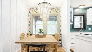 A dining area with a wooden table, wicker chairs, a chandelier, and a window with floral wallpaper.