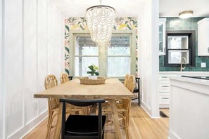 Un coin repas avec une table en bois, des chaises en osier, un lustre et une fenêtre ornée d’un papier peint à motifs floraux.