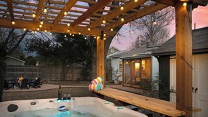 A hot tub under a wooden pergola with string lights, a wooden deck, and a house in the background.