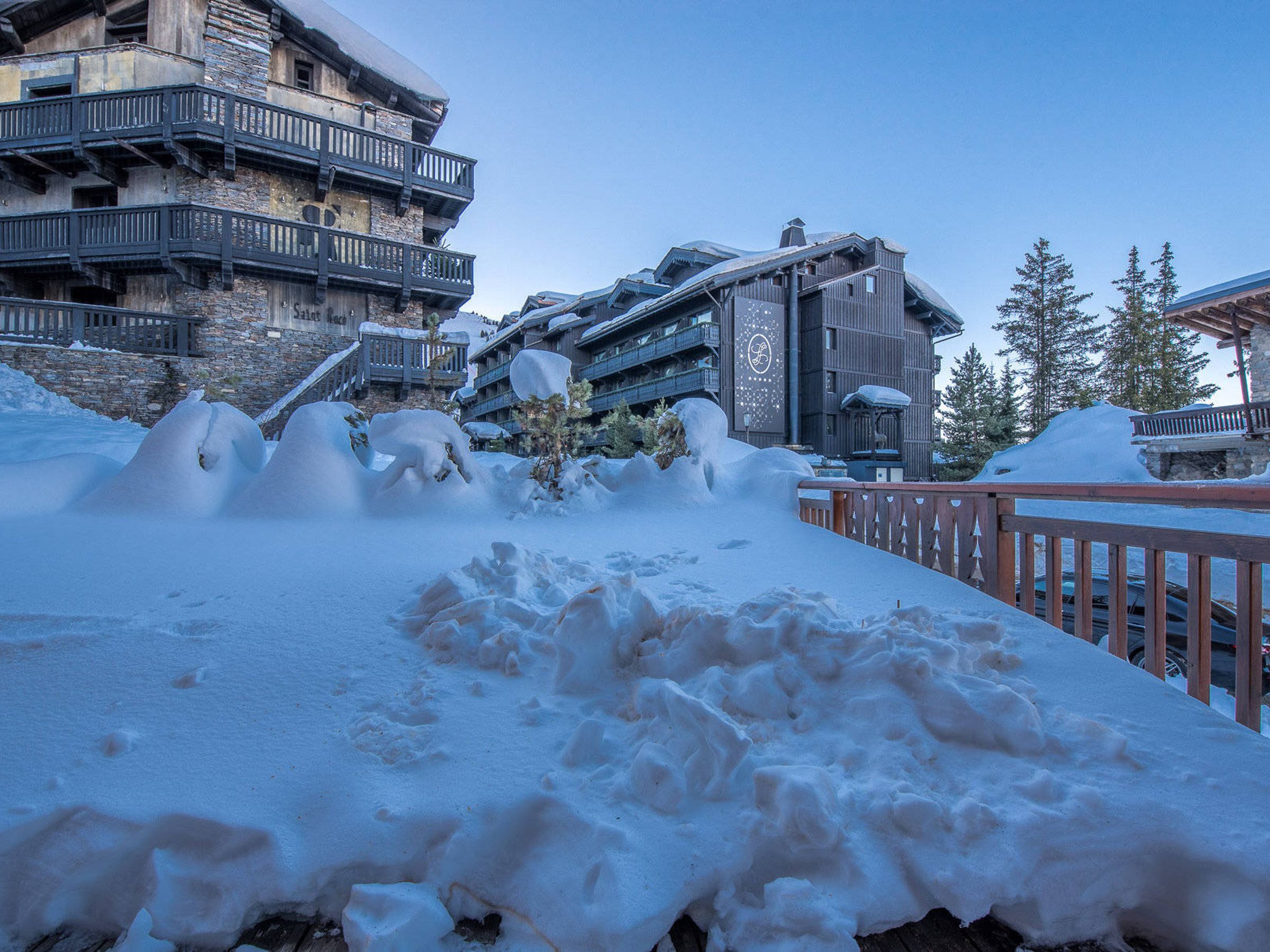 A snow-covered outdoor area with a wooden fence, a stone building with balconies, and a modern building with a ski lift in the background.