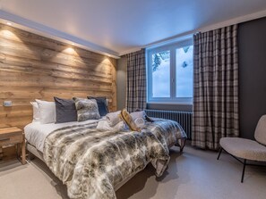 Ein Schlafzimmer mit einem Holzkopfteil, einem großen Bett mit Felldecke, einem Sessel und einem Fenster mit Vorhängen.